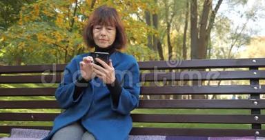 年长的女人坐在一个舒适的公园里，在智能<strong>手机</strong>里和朋友聊天。 快乐的老妇人<strong>看</strong>着屏幕